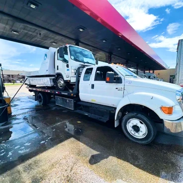 Flatbed tow truck carrying a white commercial truck at a service station, showing professional vehicle transport and roadside towing work.