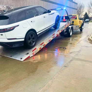 White SUV positioned on a flatbed tow truck on a wet roadway, showing vehicle transport during roadside towing operations.