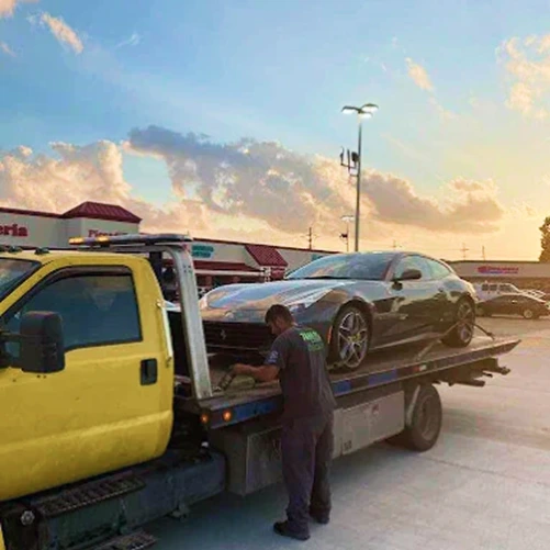 Technician securing a sports car on a flatbed tow truck in a parking lot during vehicle transport.